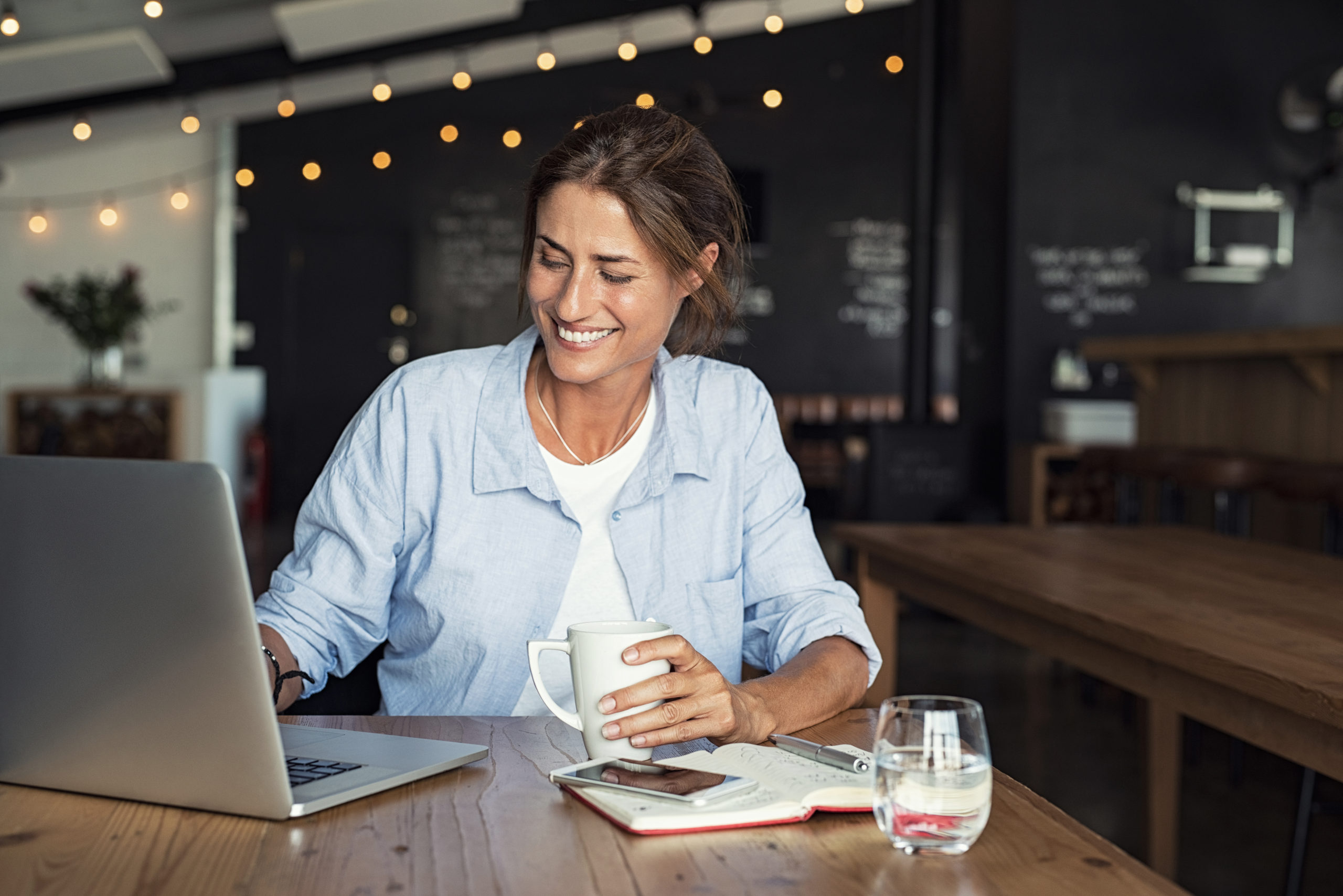 Smiling woman sitting in cafeteria holding coffee mug and working on laptop. Businesswoman checking email on laptop. Beautiful middle aged woman smiling and using laptop at cafe while drinking a cup of tea.
